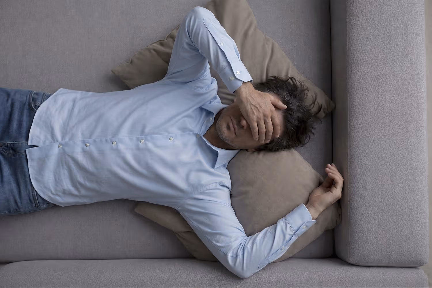Erschöpfter Mann liegt auf einem Sofa und bedeckt seine Augen mit der Hand, während seine angespannte Körperhaltung Müdigkeit, mentale Belastung und emotionale Erschöpfung ausdrückt.