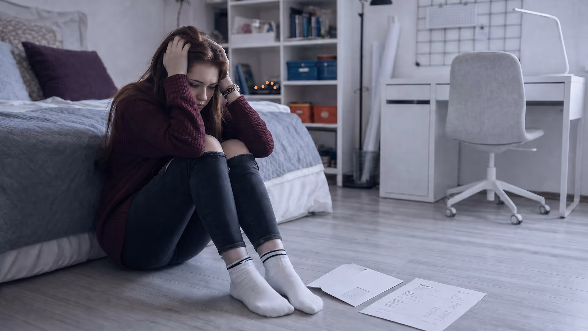 Junge Frau sitzt zusammengesunken auf dem Boden ihres Schlafzimmers und hält ihren Kopf mit beiden Händen fest, während ihre Körperhaltung Gefühle von Stress, Überforderung und emotionaler Belastung vermittelt.