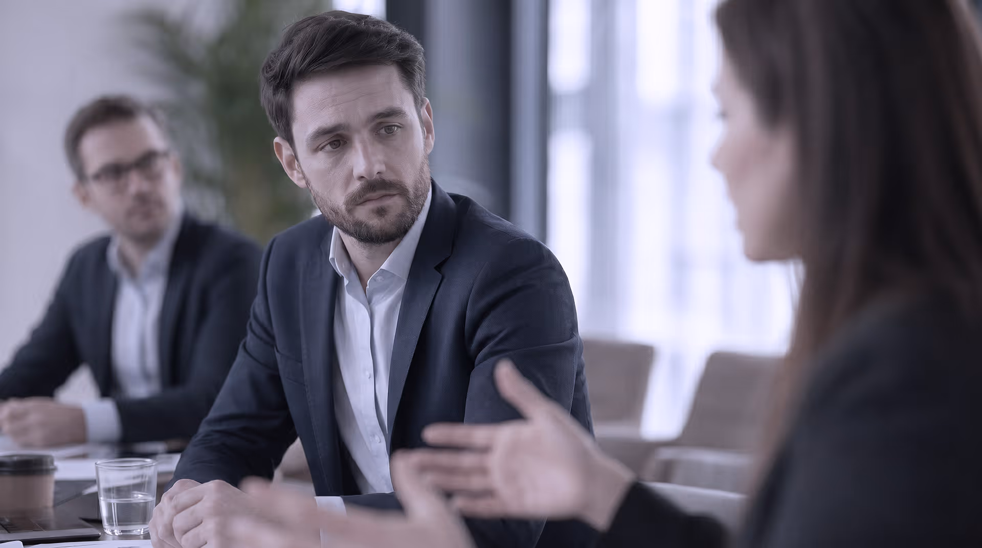Nachdenklicher Mann im Business-Outfit sitzt in einem Meeting und hört aufmerksam zu, während die angespannte Gesprächssituation Unsicherheit, Selbstzweifel und emotionale Anspannung vermittelt.
